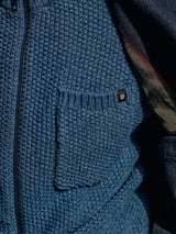 Close-up of the Aimone Knit Cardigan in Sugar Blue by Blue de GĂȘnes. Made of lambswool in coarse pearl knit, it features a front pocket with a small black logo patch displaying a face design and a partially visible zipper.