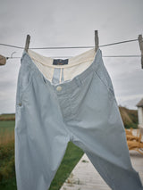 Palto Riga Chino - Blue Stripe by Blue de Gênes hang on a clothesline against a cloudy sky, secured by wooden pegs. Their relaxed fit and button fly contribute to a casual style, with lush green fields and a rustic wooden boardwalk in the background.