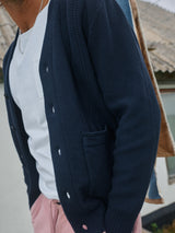 A man wears the Blue de Gênes Achille Cardigan in navy over a white t-shirt, paired with pink pants. A necklace peeks out as a blurred building with a rooftop is visible in the background.