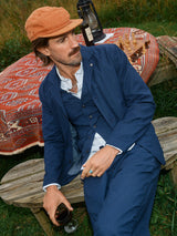 A man in a navy Ludovico Tarlo Gilet by Blue de Gênes and an orange cap sits on a wooden bench holding red wine. Behind him, an outdoor table with a patterned tablecloth, chessboard, and lantern enhances the grassy, relaxed scene.