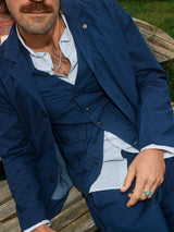 Sporting a mustache, accessorized with necklaces and a ring on their left hand, the person sits stylishly on a wooden bench in front of a grassy backdrop, wearing the Ludovico Tarlo Gilet - Navy by Blue de Gênes and donning their signature flag pin.