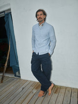 A bearded man with long hair leans barefoot against a wall on a wooden deck, wearing a light gray button-up shirt and Blue de Gênes Mustang Cargo Pant in navy. A blue curtain is partially visible on the left.