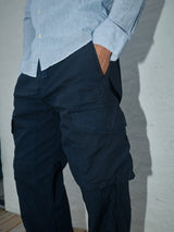 A person wearing a light blue striped button-up shirt and Blue de Gênes Mustang Cargo Pant in navy, made from parachute cotton, stands against a white textured wall. Their hands are in the utility pockets, highlighting the outfit from the waist down in the photo.