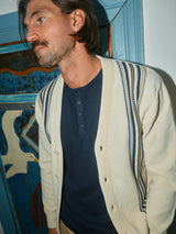 A dark-haired man with a mustache wears the Blue de Gênes Cappa Stripe Cardigan in whitecap over a navy shirt, standing before a painted wooden door featuring bird designs.
