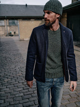 A bearded man in a Blue de Gênes Oscar Melange Beanie - Forest and a dark blazer stands on a cobblestone street by a brick building. His gray sweater, made from a cozy wool blend, complements the urban, overcast setting as he looks to the side with one hand in his jeans pocket.