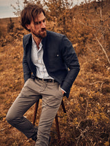 A bearded man sits on a wooden stool in a field with dry autumnal foliage, wearing a Blue de Gênes Casella Officers Blazer - Dark Navy over a striped shirt and light trousers. The setting sun casts warm light, enhancing the natural, rustic scene with vintage parade uniform charm.