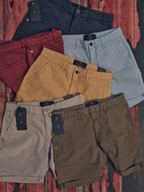 Six pairs of Teo Milano Shorts by Blue de Gênes, in navy, red, light gray, mustard, brown, and olive colors, are arranged on a worn red wooden surface. Each cotton/linen blend pair features a tag near the waistband.