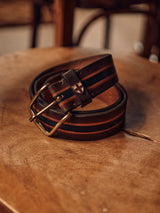 The Tino Belt - Brown Stripe by Blue de Gênes, featuring a coiled design with black stripes and a metal buckle, rests on a wooden surface. Handmade in Italy, it includes a shield emblem with a red cross and boasts a vegetable-tanned finish enhanced by the subtle background blur.