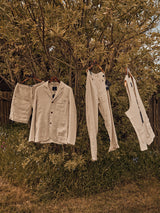 Three beige garments, including the Gabbiano Winnot Blazer - Natural by Blue de Gênes made of cotton linen, hang on hangers from tree branches. Accompanied by pants and a sleeveless top, this outdoor scene features grass and a wooden fence in the background with a warm, rustic tone.