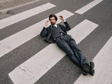 A man in a Panco Dori Chino - Grey stripe suit by Blue de Gênes lies on a zebra crossing, the crosswalks lines forming an irregular pinstripe pattern against the asphalt. His relaxed pose suggests hes resting or deep in thought.