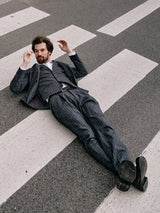 Wearing a sleek Blue de Gênes Leopardi Dori Blazer - Grey Stripe, a man lays casually on an empty zebra crossing, gazing up with relaxed elegance. His arms are raised slightly and legs crossed, showcasing the refined touch of Portuguese tailoring.
