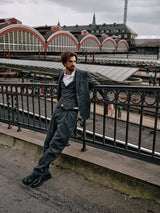 A bearded man in a Blue de Gênes Panco Dori Chino grey stripe suit leans against a bridge railing. An arched-roof train station is in the background under an overcast sky.