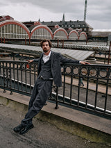 A man in the Leopardi Dori Blazer in grey stripe by Blue de Gênes leans against a black metal railing in an urban setting. Behind him is a large building with arched windows and a roofed walkway under the overcast sky.