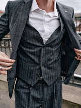 Wearing Blue de Gênes’ Canna Dori Gilet in gray stripe, a person adjusts their jacket to reveal elegant Portuguese tailoring beneath a white shirt. The outdoor photo with a metal railing highlights the sophistication of the Italian cotton-wool blend ensemble.