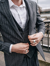 A person in a Canna Dori Gilet - Grey stripe by Blue de Gênes adjusts their jacket, showcasing Portuguese tailoring. Standing outdoors by a railing under an overcast sky, silver rings adorn their fingers, adding elegance to the Italian cotton-wool blend ensemble.