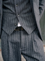 A person wearing the Blue de Gênes Panco Dori Chino - Grey stripe suit with an irregular pinstripe pattern and a visible black belt, buttoned at the waist over a white shirt, set against a blurred dark green backdrop.