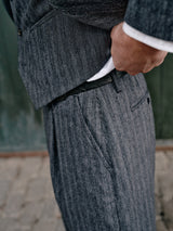Close-up of a person wearing a Blue de Gênes Panco Dori Chino in gray stripe Italian cotton-wool blend, adjusting their trousers. The suit jacket is partially visible over a white shirt as they stand on a cobblestone surface.