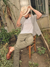 A man sits outdoors on a wooden chair, sporting the Blue de Gênes Colonnello Delo shirt in a multi-pattern over a white T-shirt, with olive pants and brown loafers. He tips his beige fedora hat amid greenery and near a structure made of corrugated metal and glass.