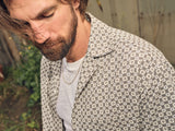 A bearded man in a Colonnello Delo shirt by Blue de Gênes, layered over a white undershirt, looks down. A chain necklace adorns his neck, partially concealed by his hair. The out-of-focus greenery enhances the Blue de Gênes aesthetic of his multi-patterned attire.