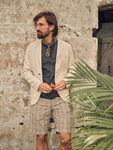 A bearded man with long hair stands against a weathered stone wall wearing a beige blazer over a Totti T-Shirt in Dark Navy from Blue de Gênes. He pairs it with plaid shorts, a dark vest, and scarf, holding something while gazing sideways. A palm leaf is visible in the foreground.