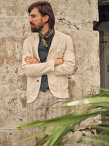A bearded man in a Blue de Gênes Gabbiano Winnot Blazer - Natural, patterned shorts, and scarf stands with crossed arms against a rustic, textured wall. Green leaves partly obscure the foreground.
