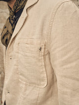 A person wears the Blue de Gênes Gabbiano Winnot Blazer in natural, a light beige cotton linen piece with a visible lapel and chest pocket adorned with a circular pin featuring a cross-like symbol. A patterned scarf is elegantly draped around the neck for added sophistication.