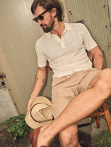 A bearded man in the Blue de Gênes Primo Polo Knit in Ecru and beige shorts sits on a wooden chair. He holds a straw hat, wears sunglasses, and poses casually against a textured wall with greenery nearby—a true summer essential.