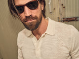 A bearded man in sunglasses dons the Blue de Gênes Primo Polo Knit in ecru, a cooling cotton/linen blend, standing confidently before a corrugated metal wall with slightly tousled hair.