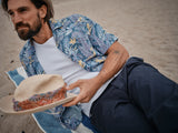 A bearded person relaxes on the beach wearing a Blue de Gênes Resort Melfi Shirt - Multi Pattern, holding a beige hat with a colorful band. They stretch out on a blue and white striped towel, surrounded by floral patterns mingling with the sandy backdrop.