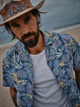 A bearded man stands on a sandy beach under a cloudy sky, wearing the Blue de Gênes Resort Melfi Shirt - Multi Pattern over a white tank top, paired with a hat featuring a colorful band.