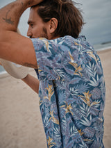 A man stands on a beach, running his hand through his hair while holding a hat. He dons a Blue de Gênes Resort Melfi Shirt - Multi Pattern, its vibrant hues contrasting beautifully against the cloudy sky and sandy shore.