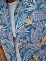 Close-up of a person wearing the Blue de Gênes Resort Melfi Shirt - Multi Pattern over a white ribbed tank top, featuring blue, purple, and yellow palm leaves with subtle flower prints. A small section of a tattoo is visible on their arm.
