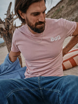 A bearded man sits on a sandy beach wearing a porcelain pink, soft cotton-jersey Salty Logo T-Shirt by Blue de Gênes, showcasing subtle chest embroidery. His blue jeans and denim jacket draped over one arm complement the scene with a striped blanket beside him and driftwood in the background.