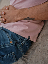 A person relaxes on the sand in a porcelain pink Salty Logo T-Shirt by Blue de Gênes and blue jeans, hands clasped over their stomach. A tattoo adorns their left arm, and a ring sparkles on their index finger, showcasing the cotton-jersey fabric and serene posture.