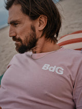 A bearded man with long hair sits on a beach, wearing the Salty Logo T-Shirt in Porcelain Pink by Blue de Gênes and a silver chain necklace. The background shows sand against a cloudy sky.