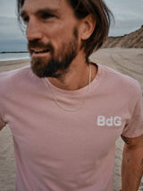 A bearded man in a Porcelain Pink Salty Logo T-Shirt by Blue de Gênes and a chain necklace stands on a beach. Sand and distant cliffs frame the cloudy sky behind him. His face is slightly blurred due to motion.