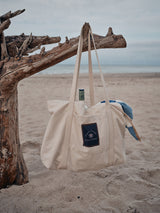 An off-white BDG Canvas Bag by Blue de Gênes hangs from driftwood on a sandy beach. Designed for daily use, it visibly holds a bottle and blue towel against a backdrop of a cloudy sky and calm sea, evoking serenity.