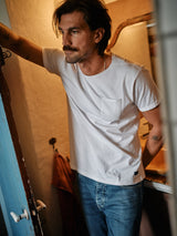 A mustached man leans casually in a doorway, wearing a Sagi Nuovo T-shirt - White by Blue de Gênes and regular fit jeans. The cozy room he stands in is warmly lit, with a bathroom seen in the background.