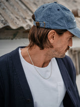 A man wearing a blue cap and an Achille Cardigan in navy from Blue de Gênes looks down. He sports a white t-shirt and a silver chain necklace. The cardigan, with its corozo buttons, stands out elegantly against the blurred, rustic roof backdrop.