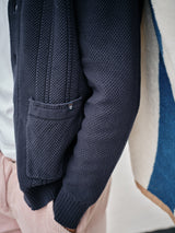 A close-up features a person in a Blue de Gênes Achille Cardigan, navy and textured, over a white shirt. The look is paired with pink pants, hands in pockets, against a blue and white backdrop.