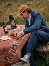 A man in Lori Tarlo Chino - Navy by Blue de Gênes plays chess alone outdoors, dressed in a relaxed-fit navy cotton outfit and cap. Hes seated on a wooden bench by a table with a red patterned cloth, surrounded by a lantern, wine bottle, and papers under an overcast sky on grass.