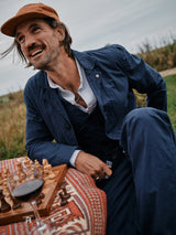 A smiling man in a Blue de Gênes Gervaso Tarlo Blazer - Navy with corozo buttons leans on a table with a chessboard and red wine glass outdoors, sitting on patterned cloth against a grassy field, perfectly blending elegance and leisure.