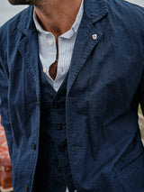 The individual sports a navy blazer adorned with a Blue de Gênes flag pin and wears a blue-and-white striped shirt. The ensemble includes the Ludovico Tarlo Gilet - Navy by Blue de Gênes, showcasing chambray fabric with buttons. The view captures their clothing details from the chest up, face partially visible.