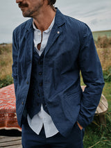A man stands outdoors in a regular fit ensemble, wearing a navy blazer over the Blue de Gênes Basso MC Shirt - Navy Stripe and a waistcoat, hands in pockets. Blurred greenery and a red-patterned rug create the background.