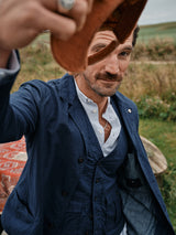 A man with a mustache, dressed in the Blue de Gênes Gervaso Tarlo Blazer in Navy and a white shirt, lifts a brown hat while standing outdoors on grassy terrain under cloudy skies.