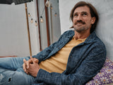 A person with shoulder-length hair and a beard is lounging outside on a patterned cushion, smiling while wearing a yellow Filo Knit Polo by Blue de Gênes over a blue shirt paired with ripped jeans. Hanging lights illuminate the backdrop.
