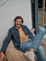 A smiling man with long hair and beard relaxes on a wicker chair in an outdoor setting near a window, wearing a yellow shirt, blue jacket, and Blue de Gênes Vinci Super Used Jeans - expertly crafted in Italy.