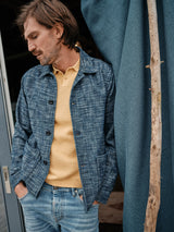 A man with shoulder-length hair leans against a wall, wearing the Blue de Gênes Kojima Jacket in navy over a yellow polo shirt and light blue jeans. He stands next to a large blue curtain and wooden pole, gazing downward with a relaxed expression.