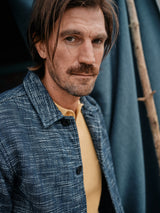 A man with medium-length hair and a mustache looks at the camera, exuding a lived-in vibe. He wears the Kojima Jacket - Navy by Blue de Gênes over a yellow shirt. A blue fabric and wooden pole in the background complete the scene.