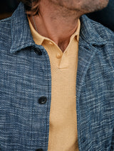 A person wearing a textured blue jacket over a sleek Filo Knit Polo in Yellow Grain by Blue de Gênes. The jacket, with visible buttons and a collar, complements the polos small notch and subtle pattern beautifully.